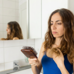 A woman in a blue tank top looks worriedly at hair on her brush, standing in a bathroom with white tiles and a mirror behind her.
