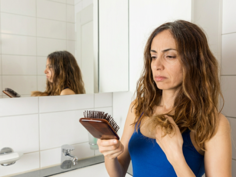 Why Is My Hair Falling Out Now? The Delayed Effects of Illness, Stress, and Weight Loss A woman in a blue tank top looks worriedly at hair on her brush, standing in a bathroom with white tiles and a mirror behind her.
