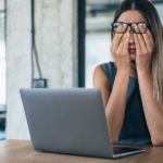 A person sitting at a desk with a laptop, covering their face with both hands, appearing stressed or overwhelmed.