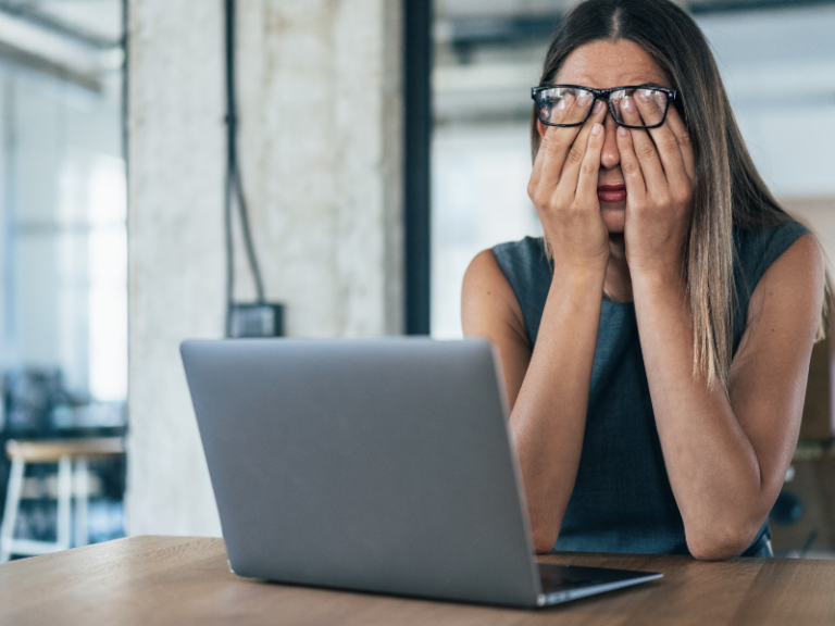 A person sitting at a desk with a laptop, covering their face with both hands, appearing stressed or overwhelmed.
