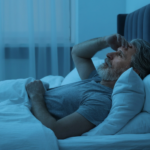An older man with gray hair and a beard lies in bed at night, looking worried or thoughtful while resting his hand on his forehead. Blue lighting suggests it is nighttime.