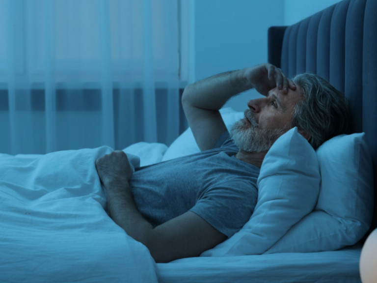 Waking Up to Urinate Again? What Nighttime Bathroom Trips May Be Trying to Tell You An older man with gray hair and a beard lies in bed at night, looking worried or thoughtful while resting his hand on his forehead. Blue lighting suggests it is nighttime.